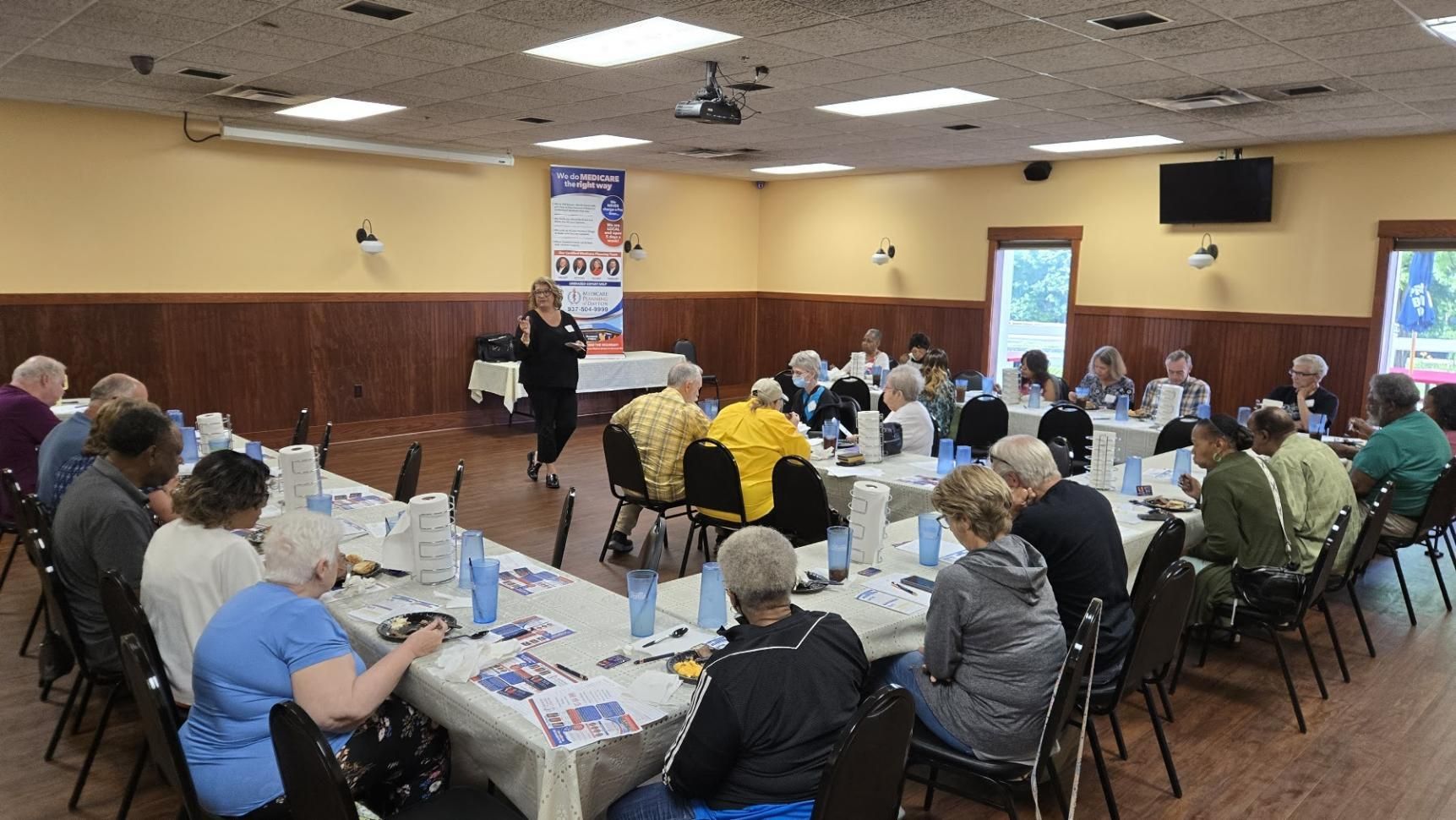 A group of people sit at long tables in a room while a speaker stands at the front .