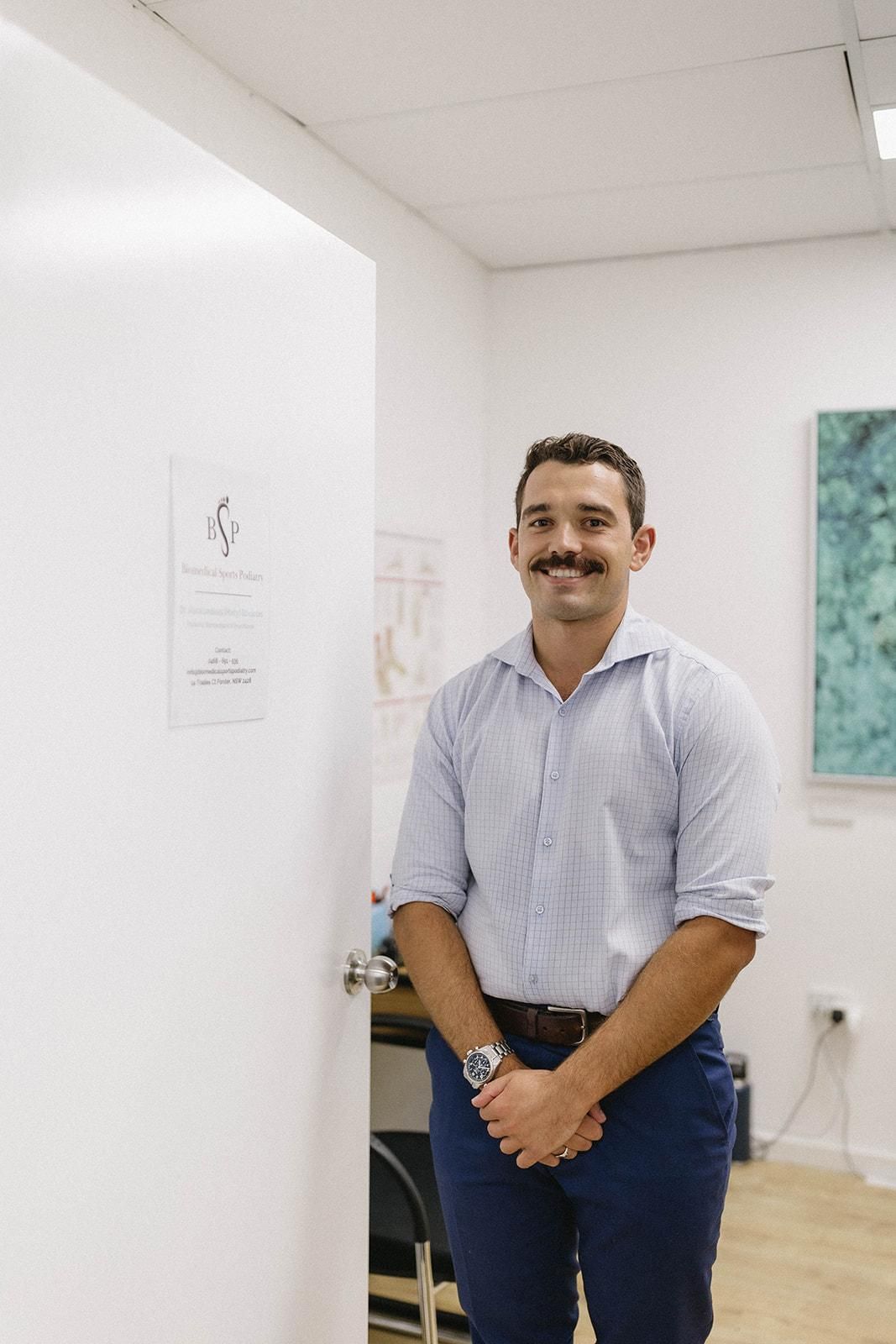 A Man With a Mustache is Standing in Front of a Door in a Room — Biomedical Sports Podiatry in Forster, NSW