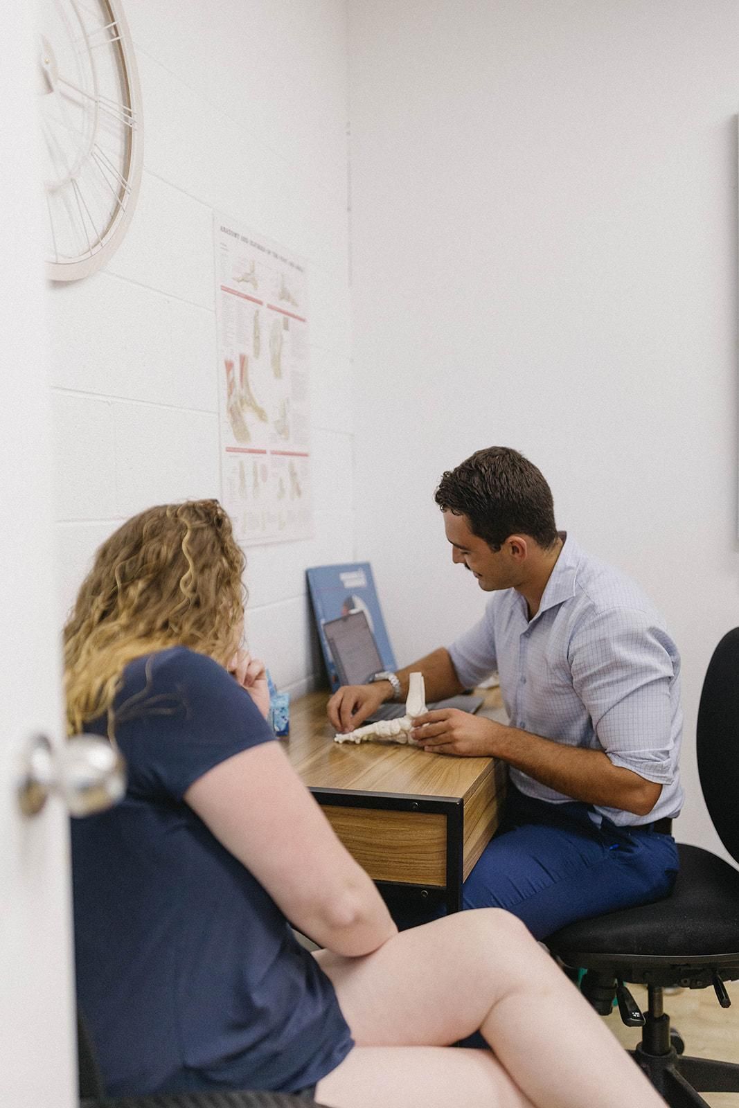 A Man is Sitting at a Desk Talking to a Woman — Biomedical Sports Podiatry in Forster, NSW