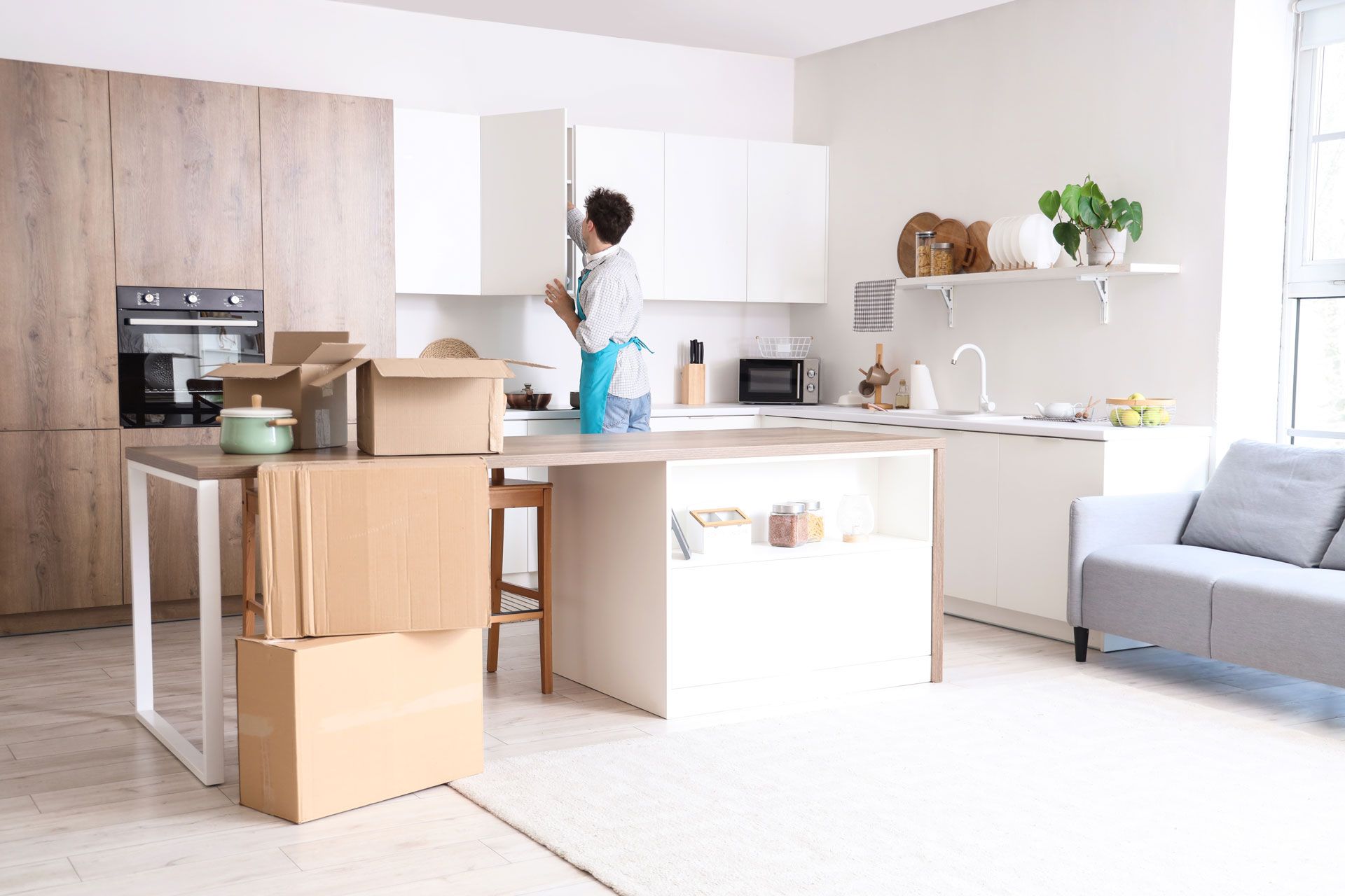 Person unpacking boxes in a bright, modern kitchen during a move. Boxes and furniture visible.