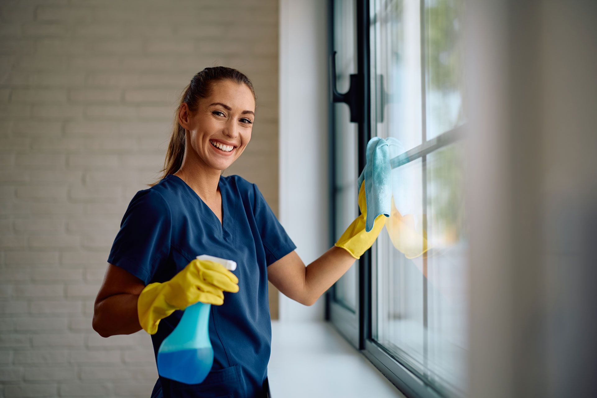 Woman in gloves cleaning a window with a smile, holding spray bottle, indoors.