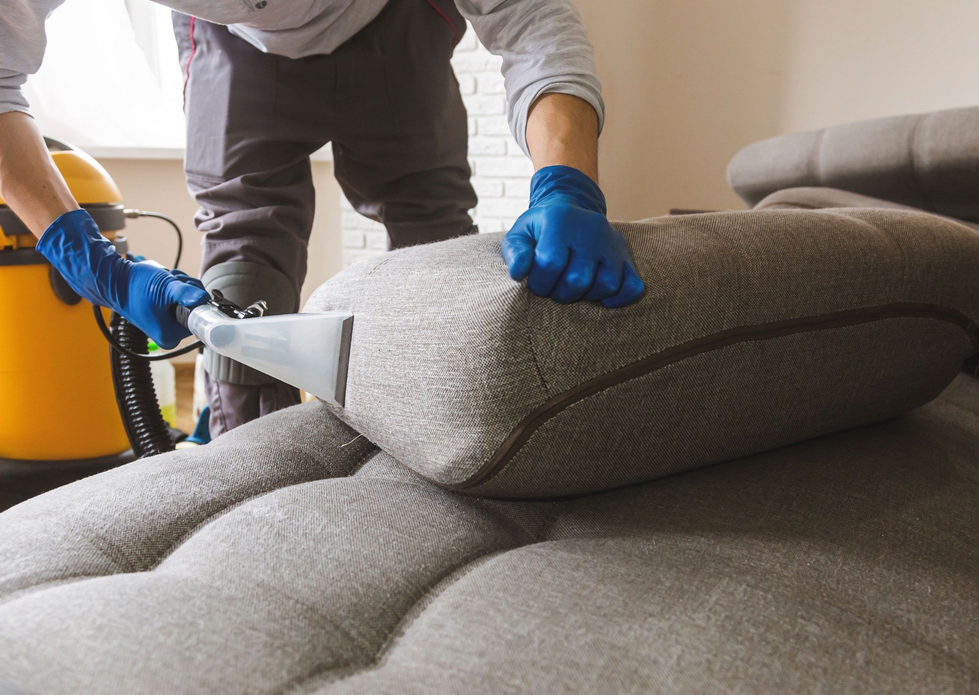 Person cleaning a gray couch cushion with a yellow upholstery cleaner, wearing blue gloves.