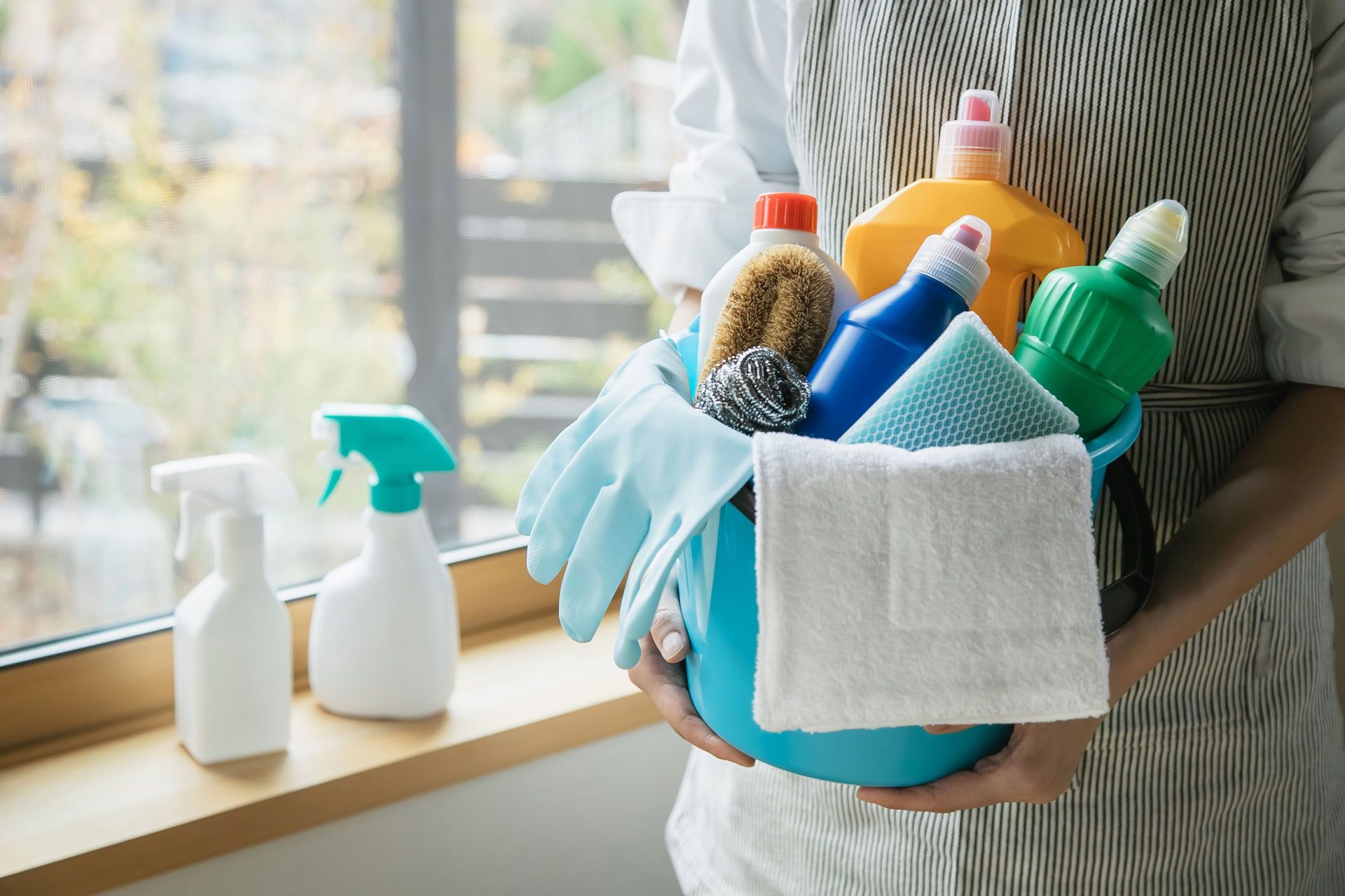 Person holding a blue bucket filled with cleaning supplies near a window.