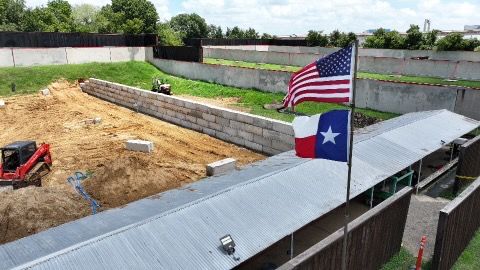 Construction site with American and Texas flags; excavator, dirt, and retaining walls visible.