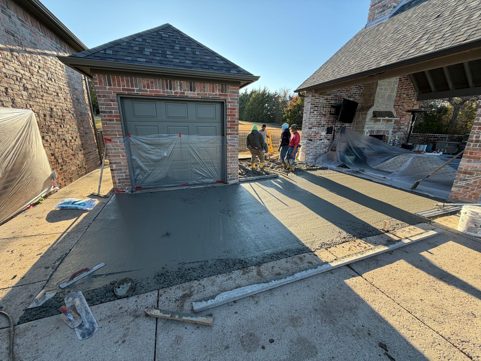 Workers pouring wet concrete for a driveway in front of a brick garage and covered patio.