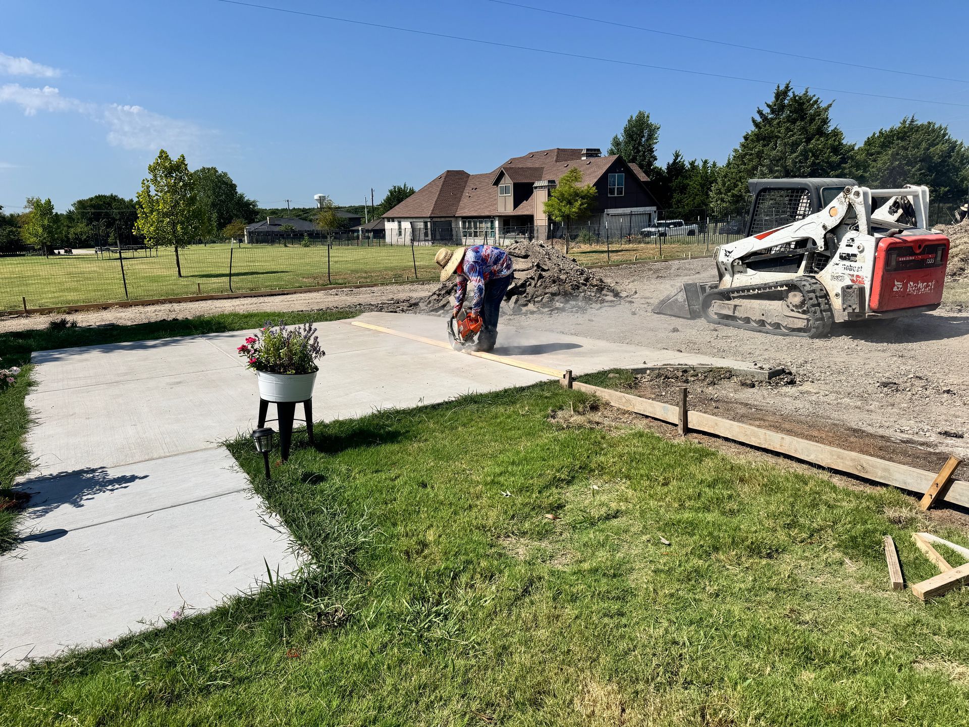 Construction site: worker cutting concrete with saw. Skid steer, grass, and house in background.