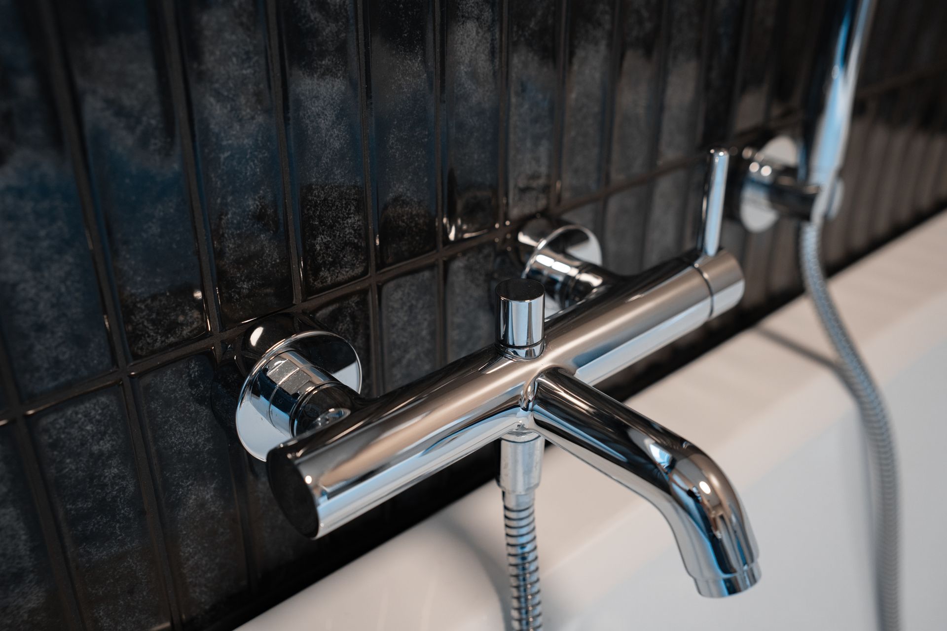 Shiny chrome faucet and showerhead on a dark-tiled wall next to a white bathtub.
