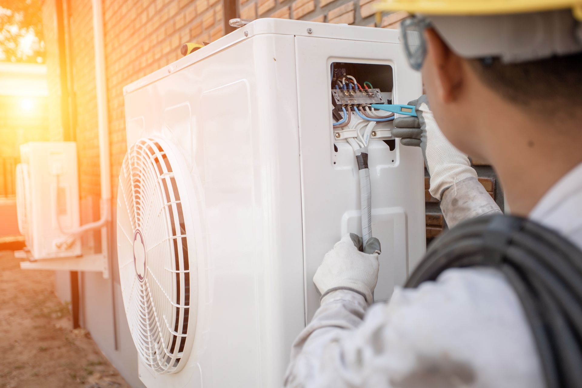 HVAC technician in a yellow hard hat repairs an air conditioner unit outdoors, sunny day.