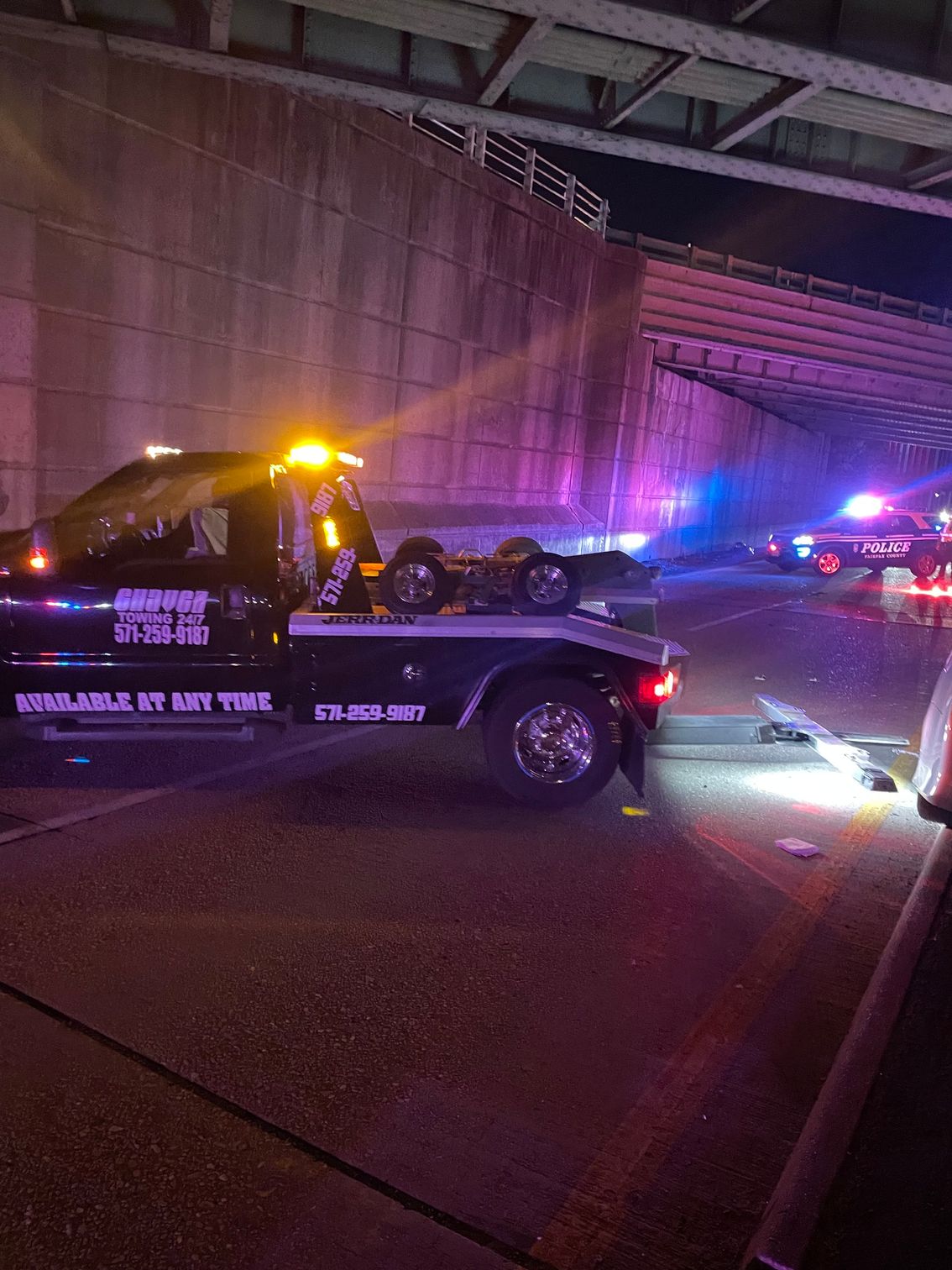 A black tow truck with flashing lights parked on a highway underpass at night, with another emergency vehicle nearby.
