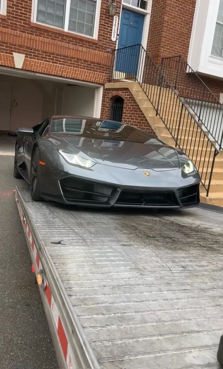 A tow truck operator in a high-visibility vest secures a white car onto the flatbed truck on an urban street.