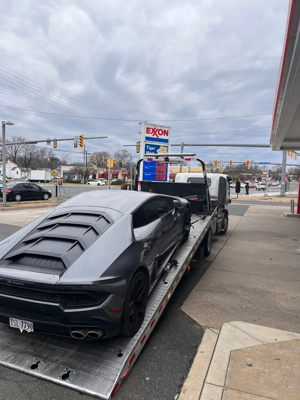 A grey Lamborghini sports car sits on a flatbed tow truck parked at an Exxon gas station under a cloudy sky.