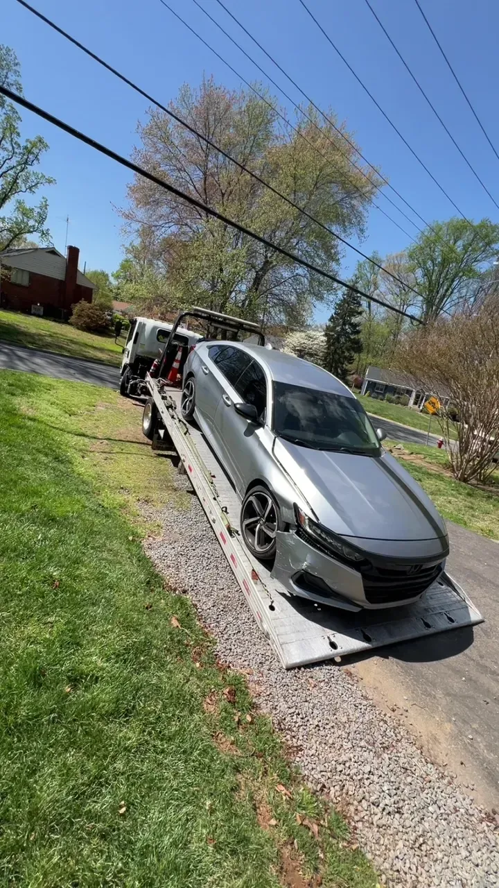 A silver sedan with front-end damage being loaded onto a flatbed tow truck parked on the side of a road.