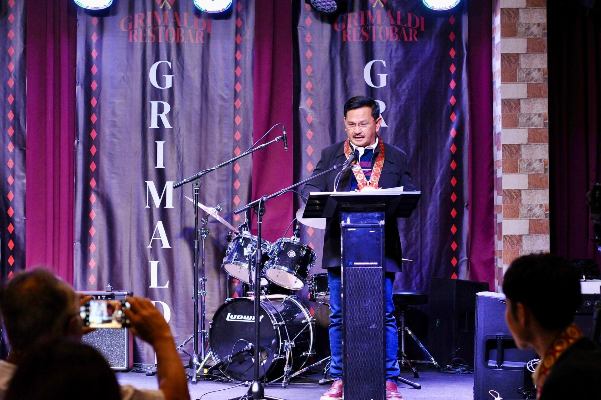 A man is standing at a podium giving a speech in front of a drum set.