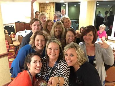 Group of smiling people pose together in a room, some waving, some smiling.  Warm lighting.