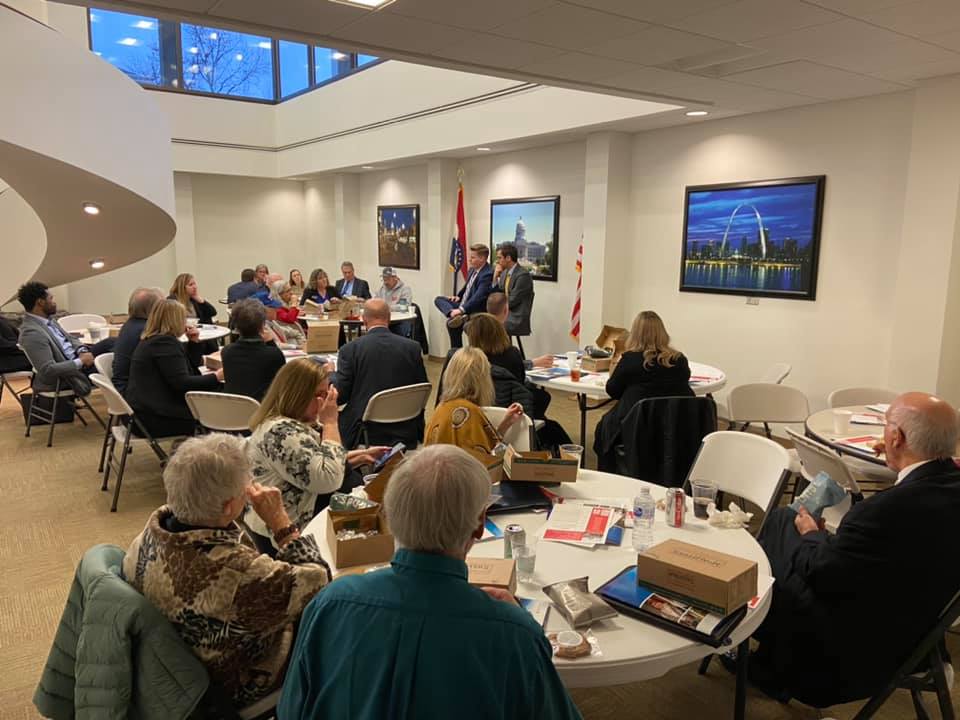 Group of people seated around tables, listening to speakers in a well-lit room.