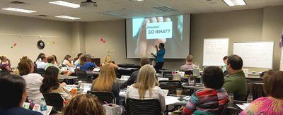 A presenter speaks in a classroom to a seated audience. A screen displays 