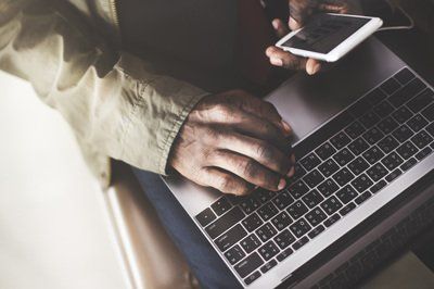Man using laptop, holding a phone. Dark lighting, indoors.