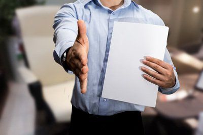 Man in a blue shirt extends hand for a handshake, holding a blank paper in the other hand.