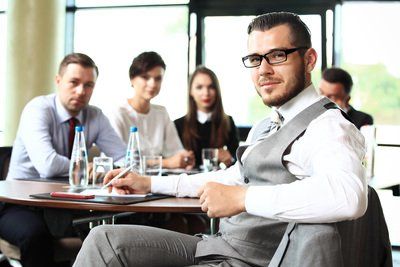Business team at a conference table, man with glasses smiling at camera, other colleagues behind.