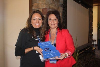 Two women holding an award in an indoor setting. One wears black, the other red, smiling.