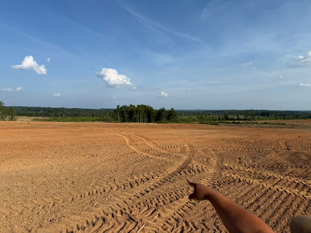 A hand points toward a wide, barren field of turned reddish-brown earth under a bright blue sky with scattered clouds.