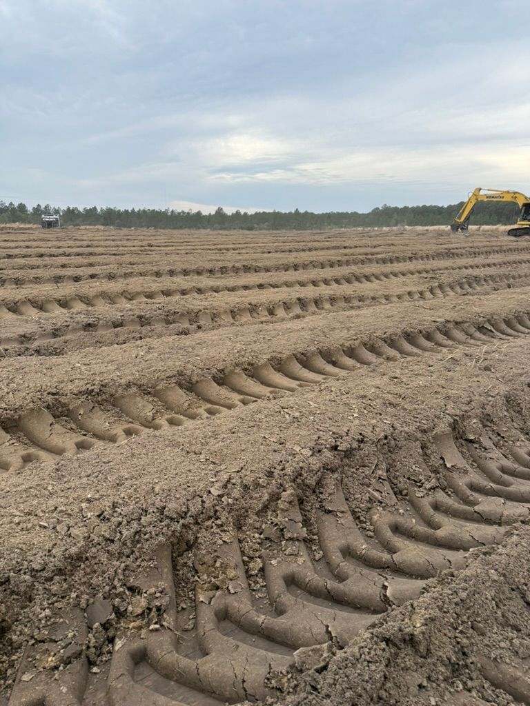 A yellow excavator sits on a vast, cleared construction site marked by deep, parallel tire tracks in the dirt.