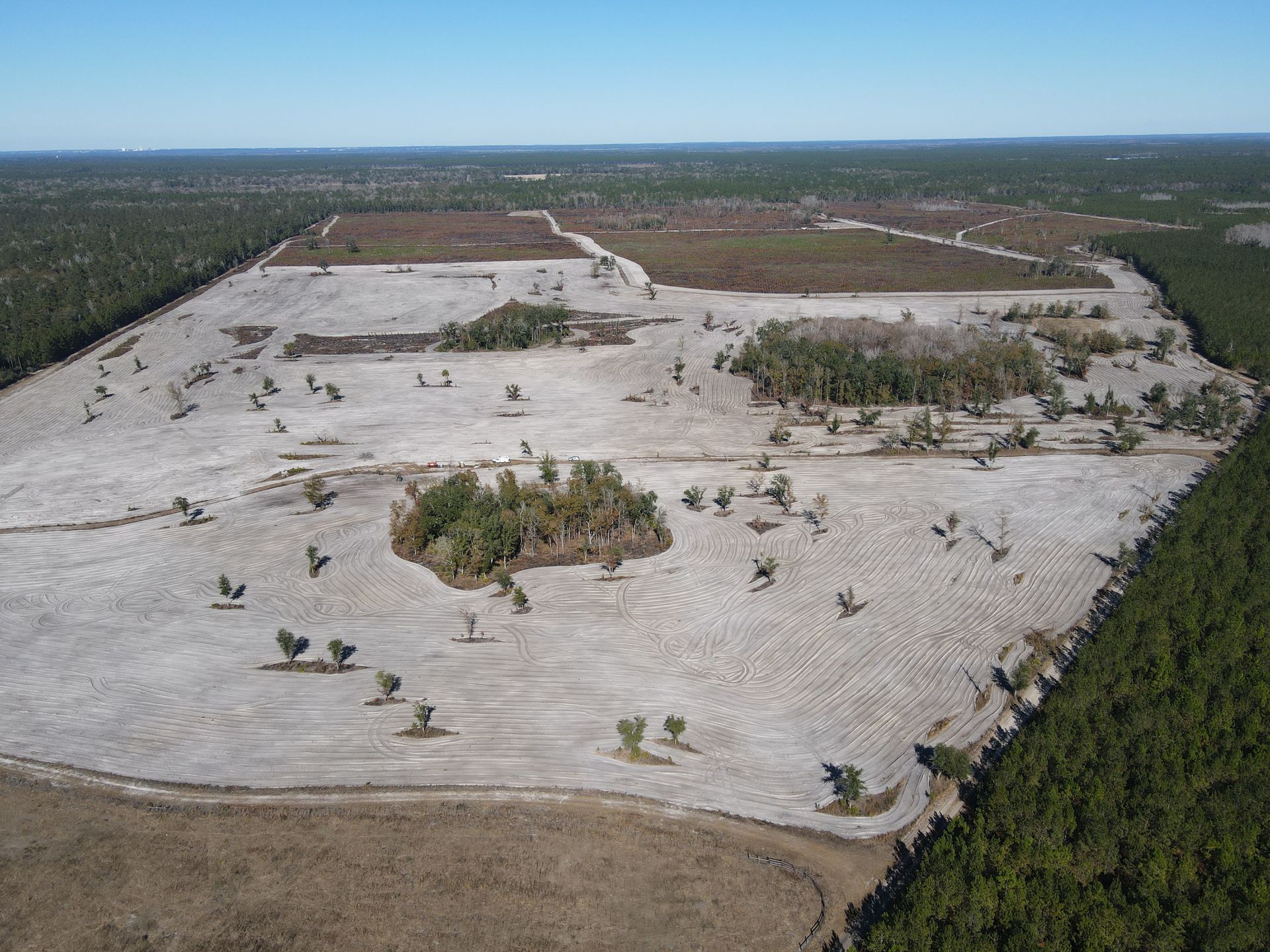 An aerial view of a large, cleared plot of land surrounded by dense pine forests under a clear blue sky.