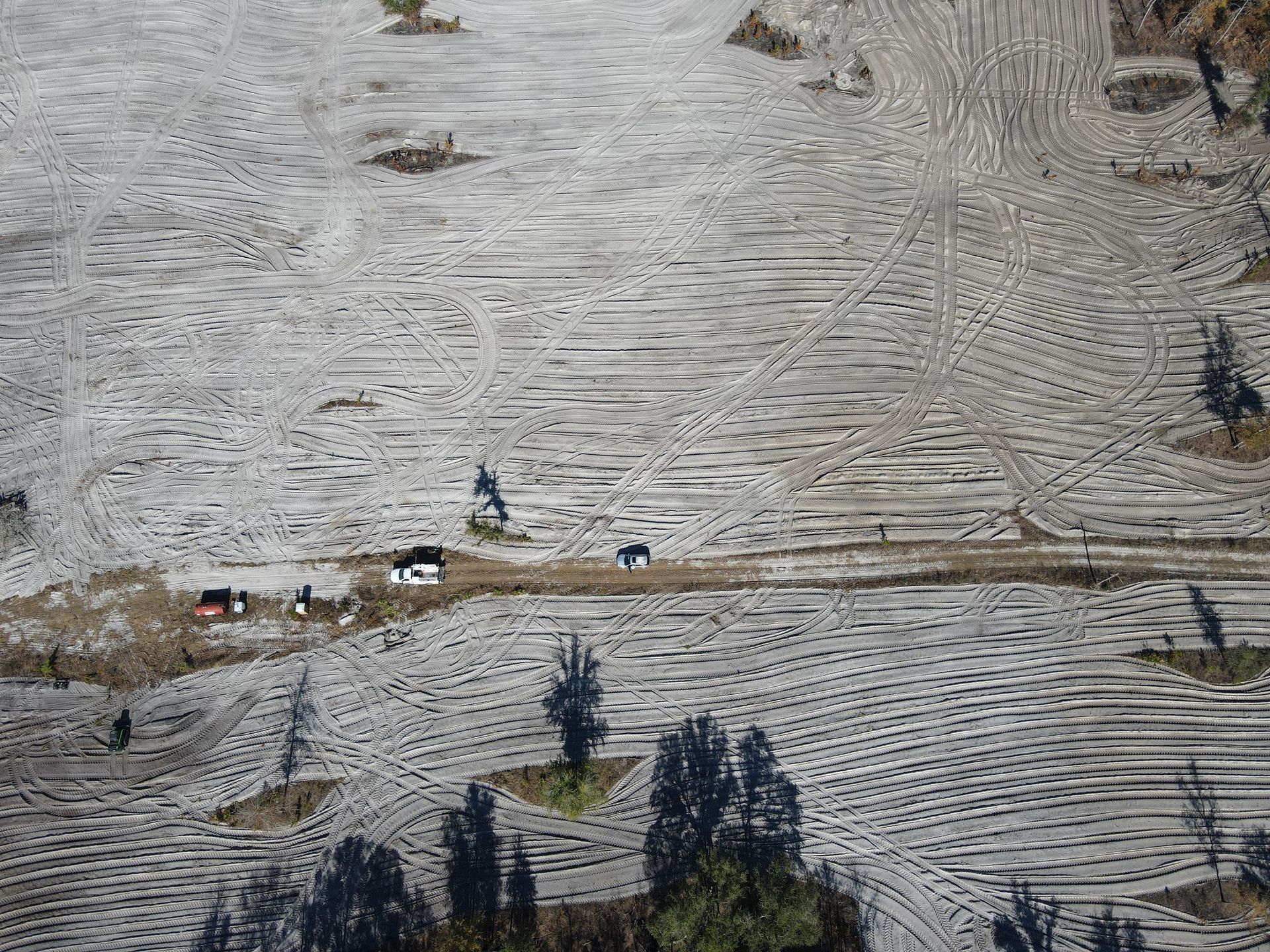 An aerial view of a barren, dusty landscape covered in rhythmic, dark furrows, with a dirt road and a few small trees.