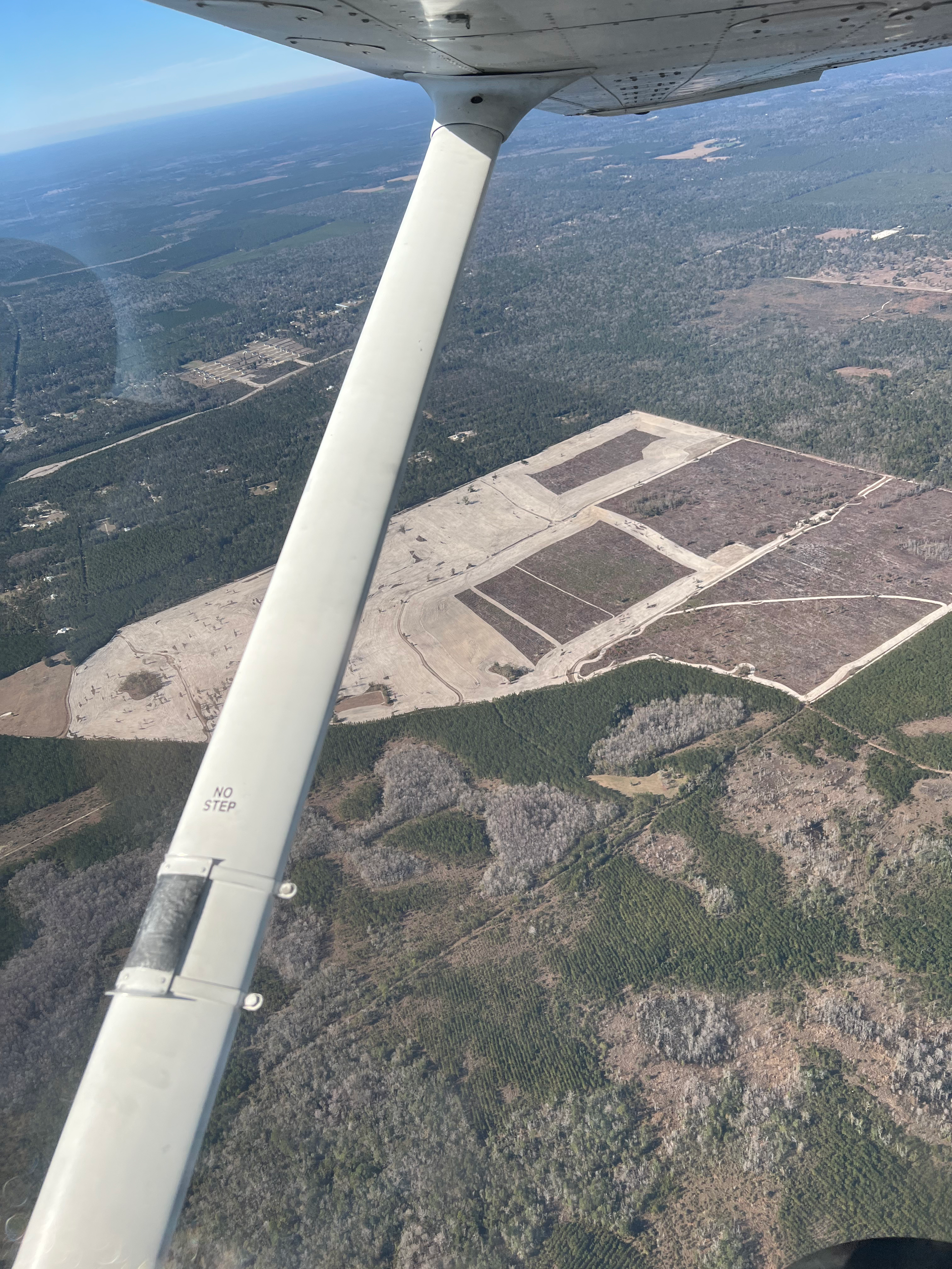 Aerial view from an airplane cockpit showing a cleared, sandy land development site surrounded by dense forest.