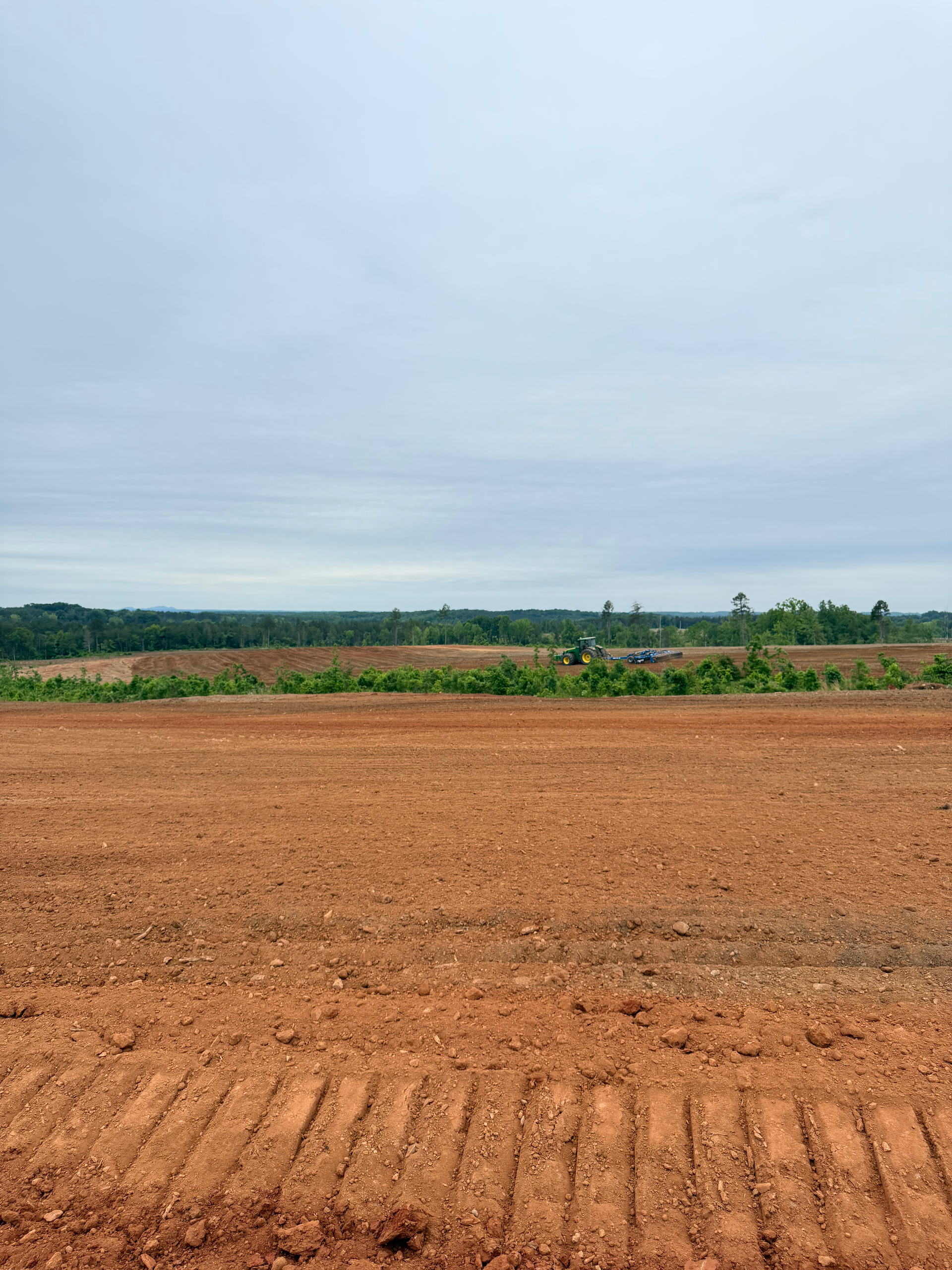A wide, freshly cleared plot of red clay soil under a cloudy sky, with a line of trees visible in the distance.