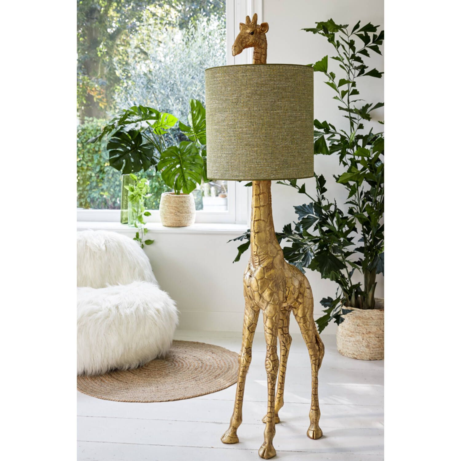 Gouden vloerlamp in de vorm van een giraffe met een groene lampenkap, in een kamer met planten en een pluizige stoel.