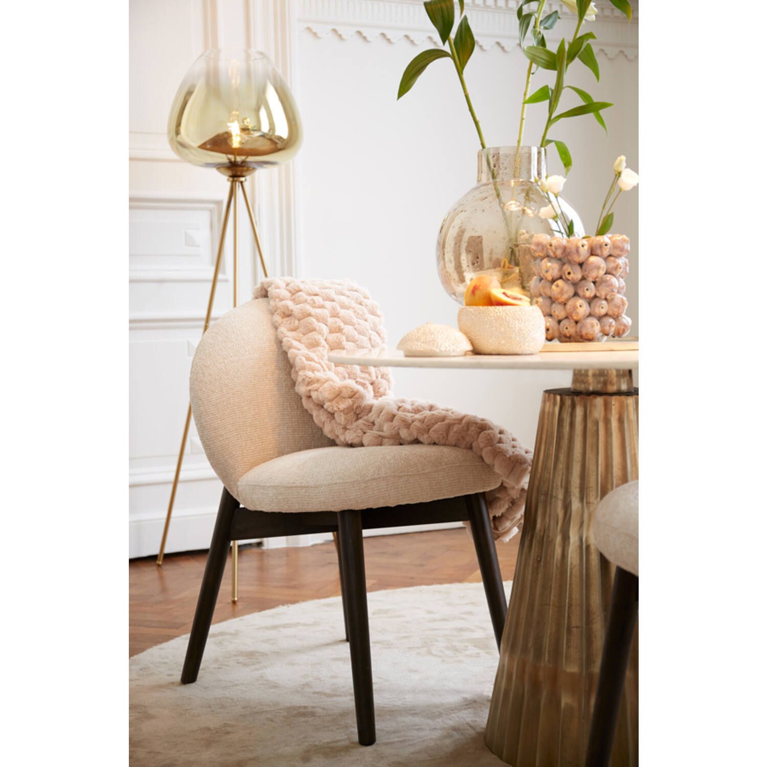 Eetkamerscène: ronde tafel met stoel, gouden accenten, glazen vaas met bloemen en een staande lamp.