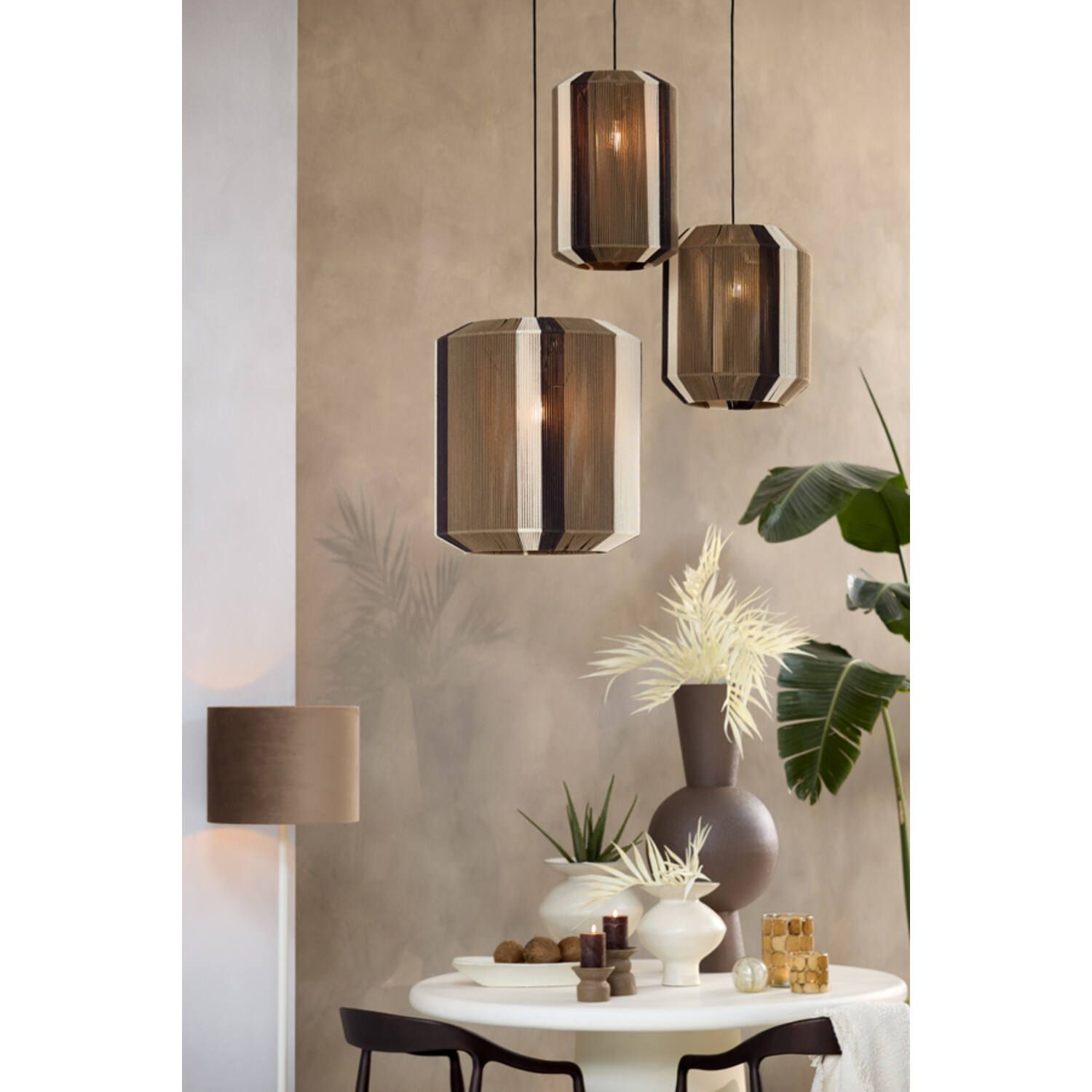 Drie hanglampen boven een eettafel. Bruine, beige en witte gestreepte kappen, met links een wandlamp en een vaas met planten.