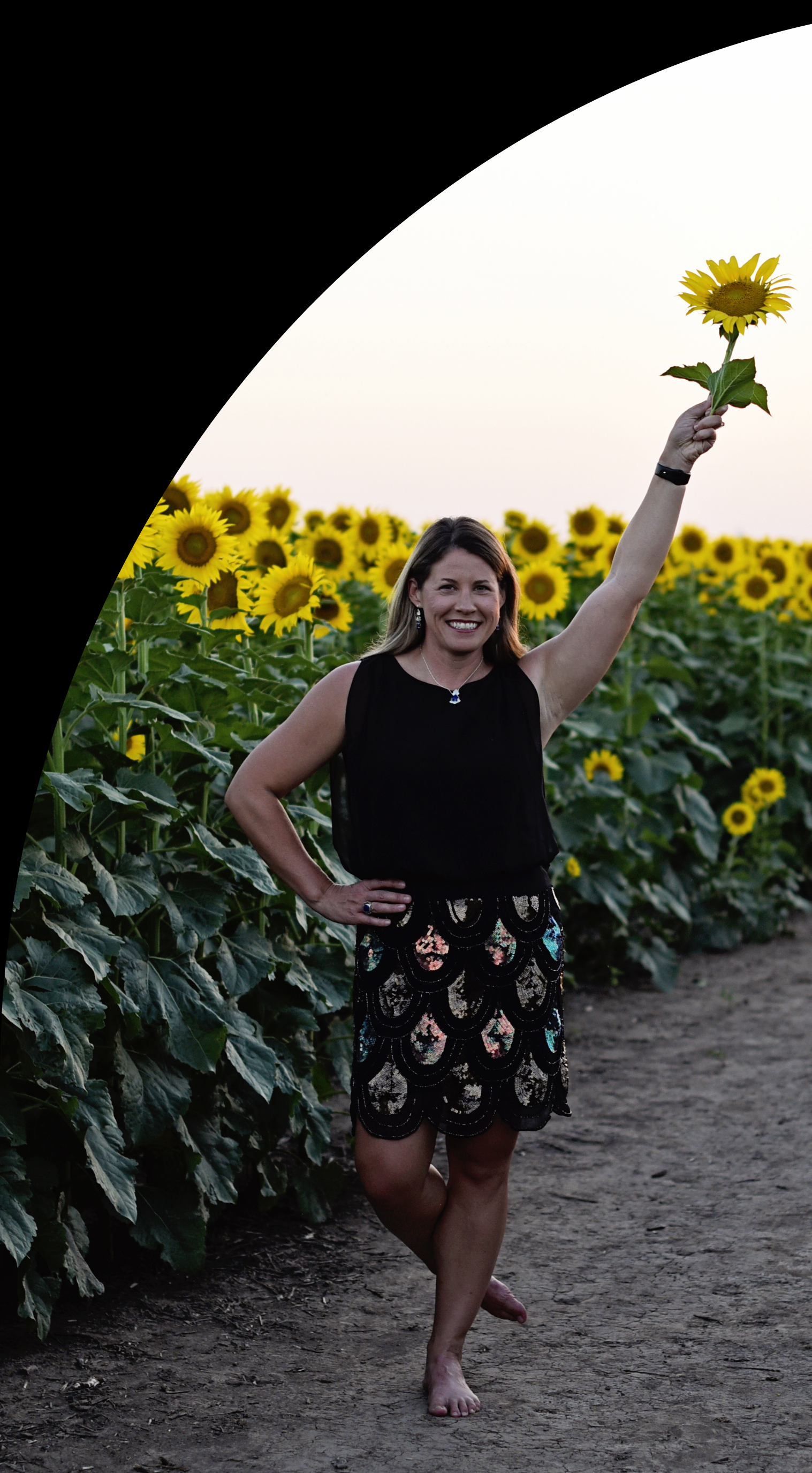 Woman in black outfit holding sunflower, smiling in a sunflower field.