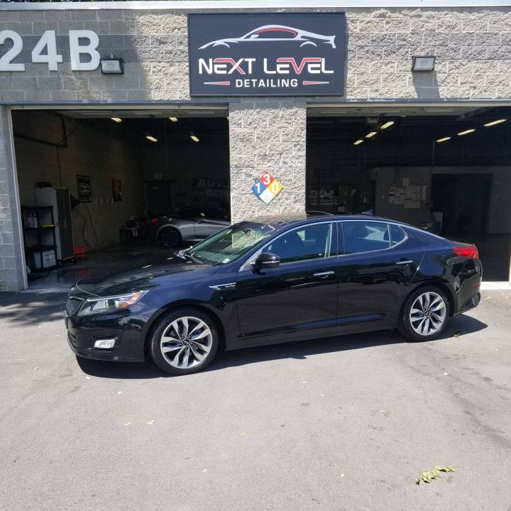 Black Kia Optima parked in front of Next Level Detailing garage with sign above.