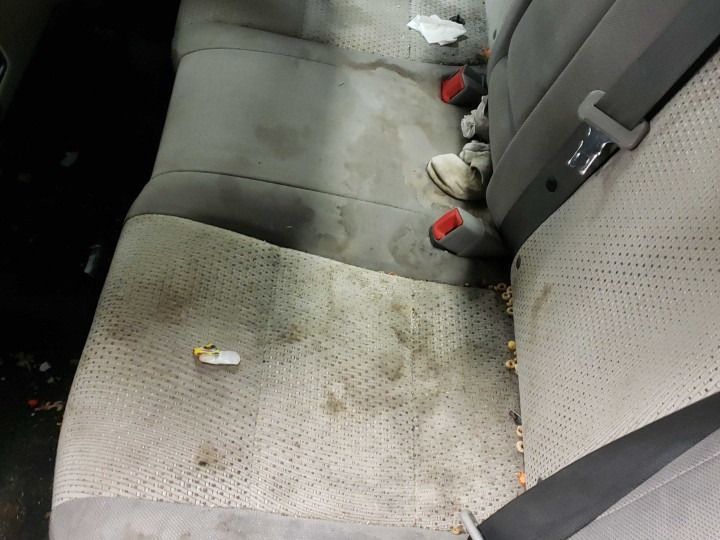 Dirty car back seat with stains and debris; tan fabric, red seatbelts.