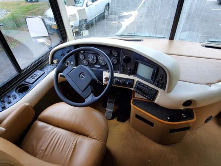 Interior view of an RV cockpit, tan leather seat, black steering wheel, gauges, and windshield.