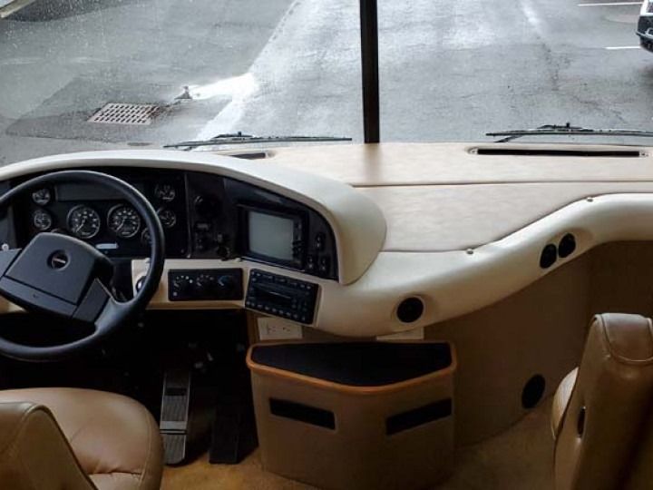 Interior view of a beige RV dashboard with steering wheel, gauges, and a small screen; street visible through the windshield.