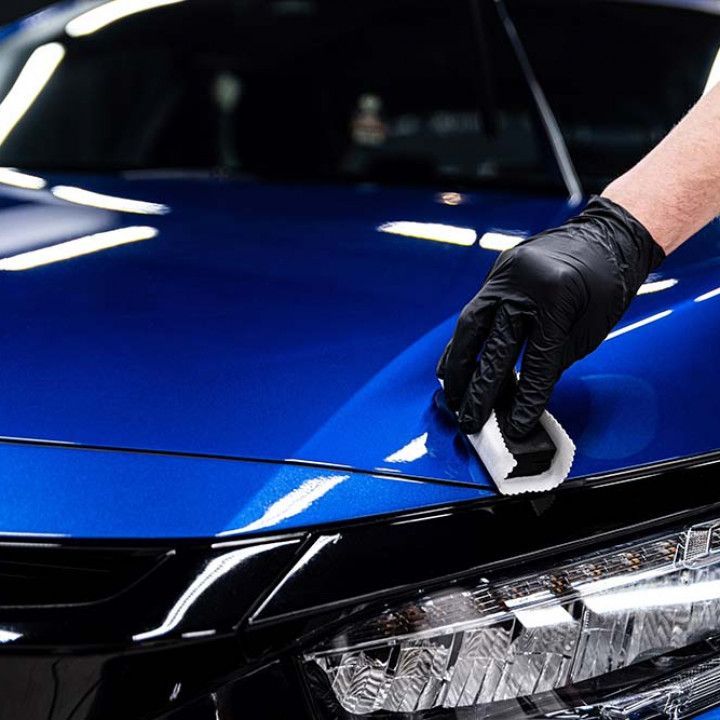Gloved hand applying ceramic coating to a blue car's hood, inside a well-lit setting.