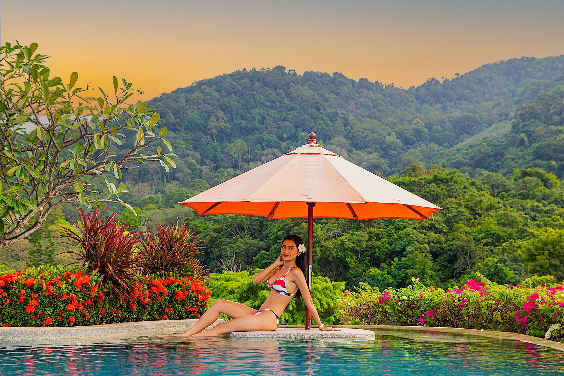 A woman in a bikini sits under an umbrella near a swimming pool