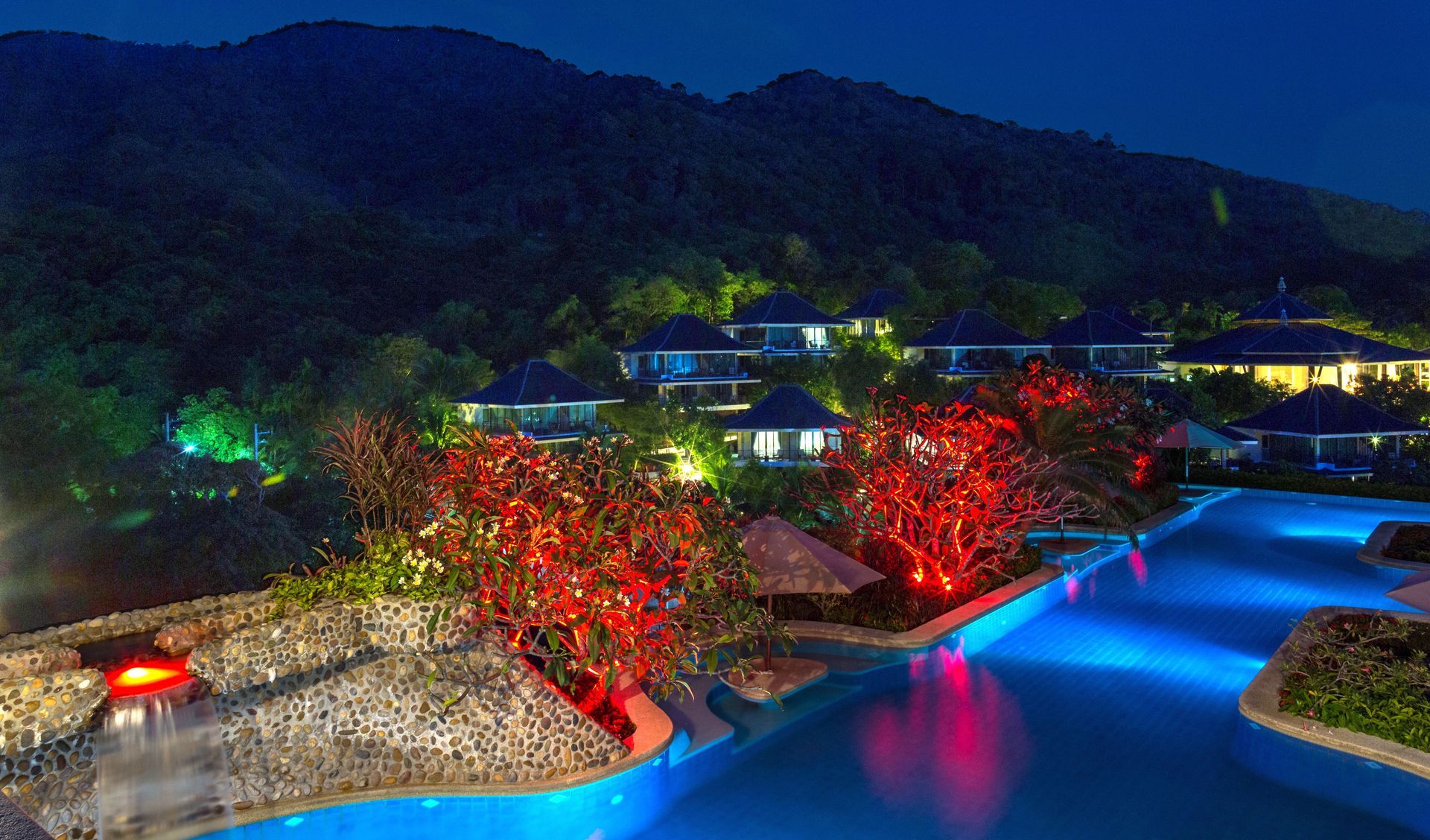 A large swimming pool is lit up at night with mountains in the background