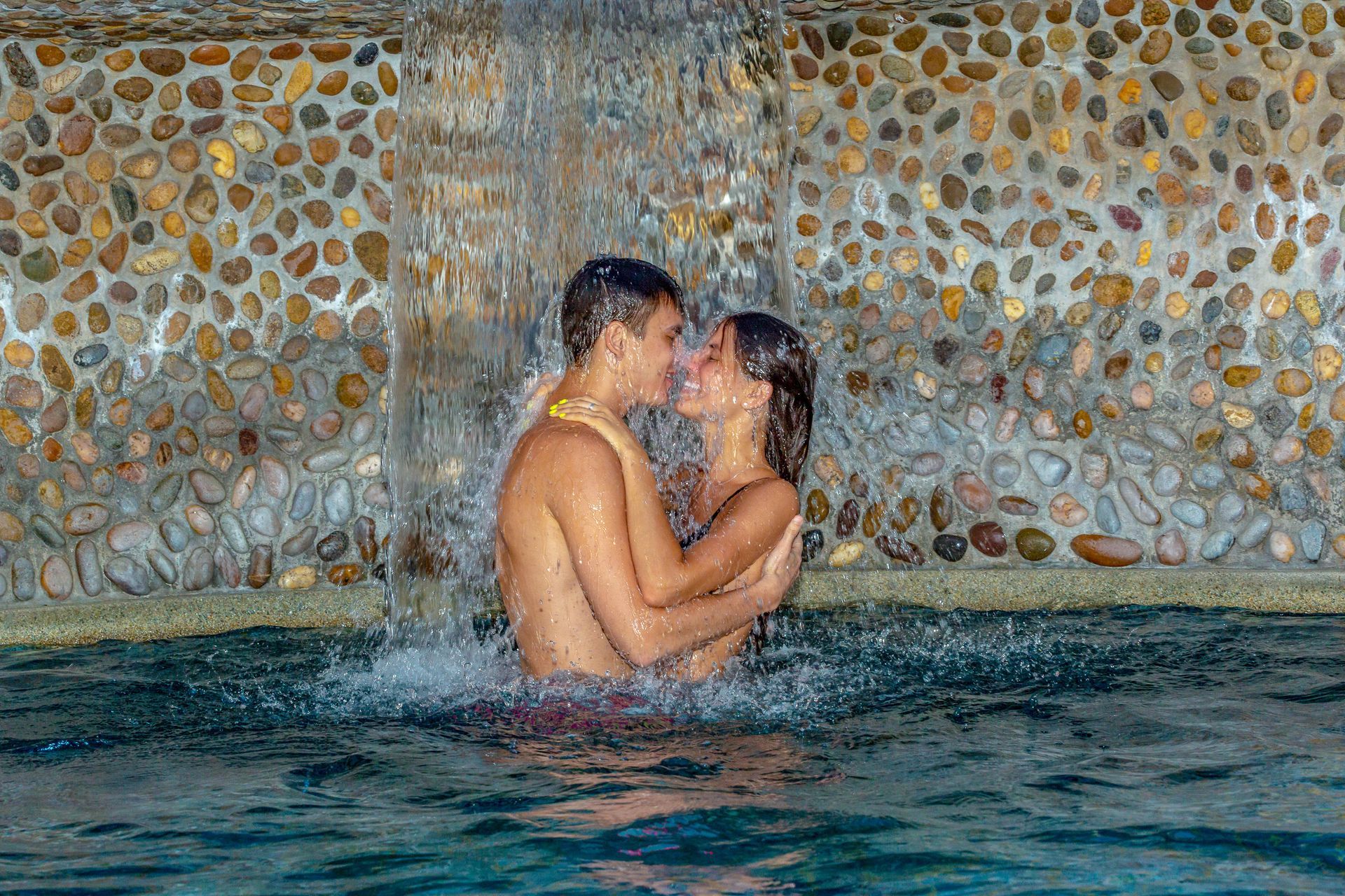 A man and a woman are kissing in a swimming pool under a waterfall.