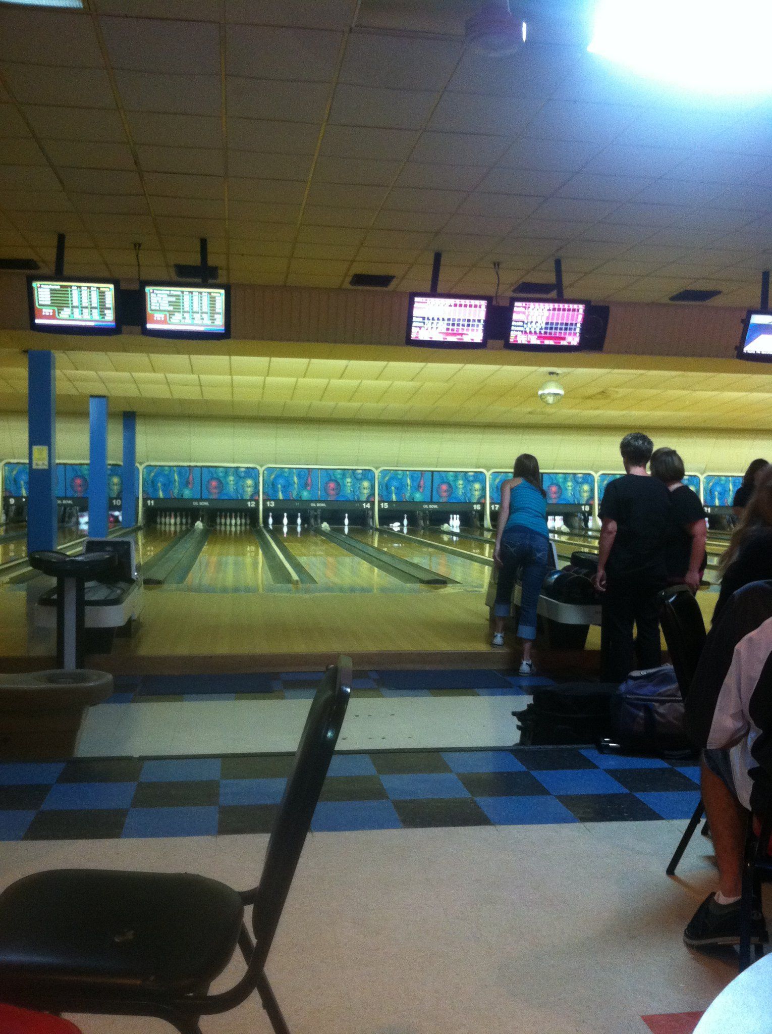Bowling alley: People standing near lanes, awaiting their turn to bowl, with television screens above.