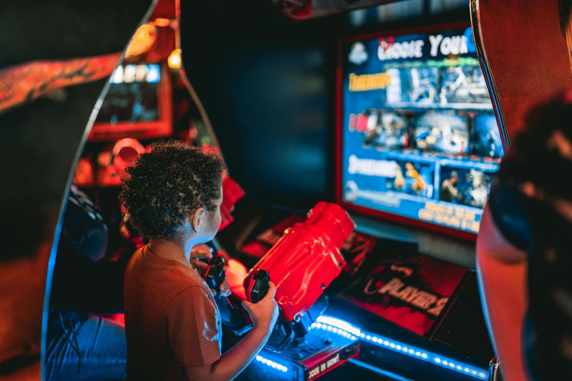 Child playing a red light gun arcade game, lit with blue neon, in a dimly lit arcade.