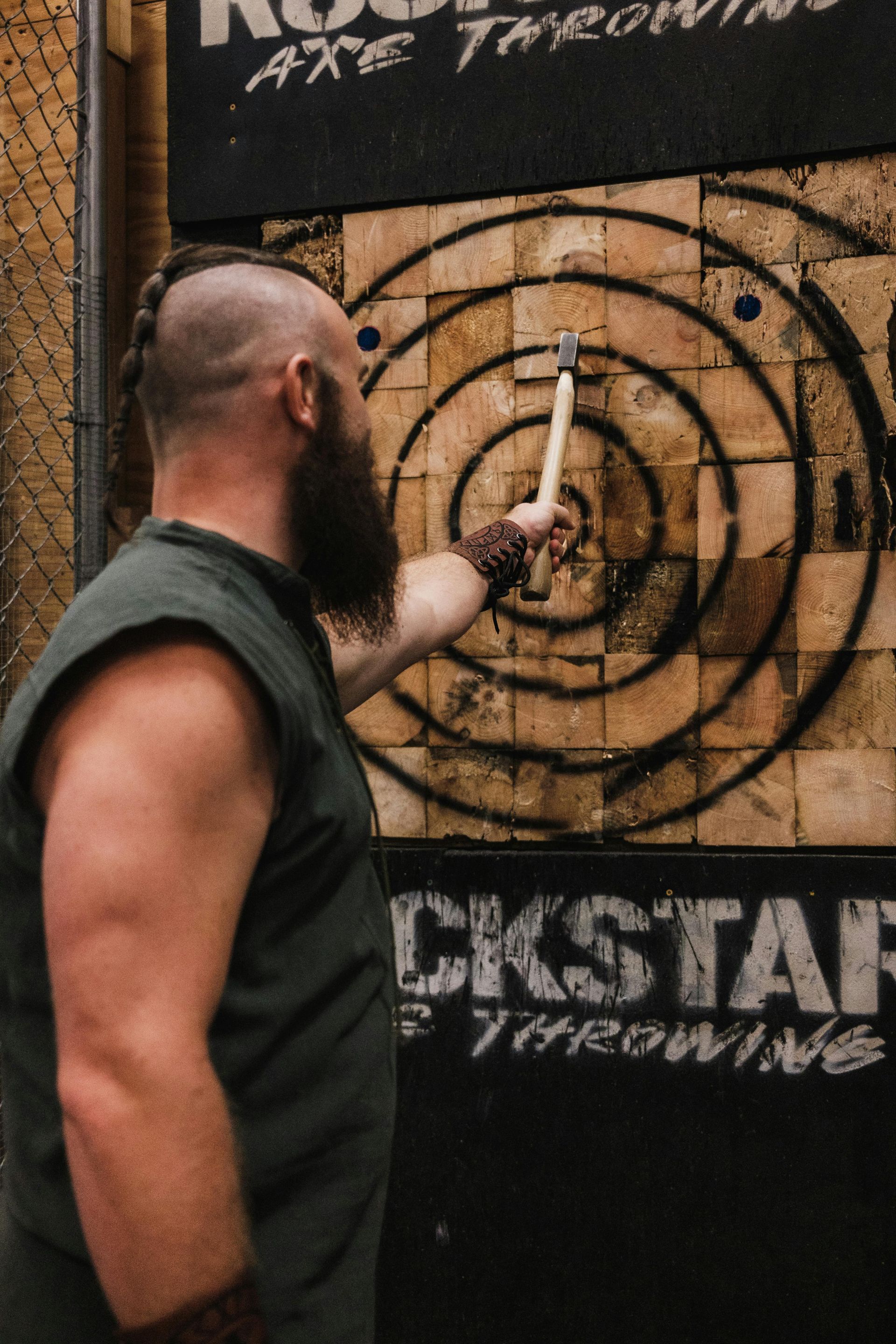 Man with beard holding an axe, pointing at a target in an axe throwing venue.