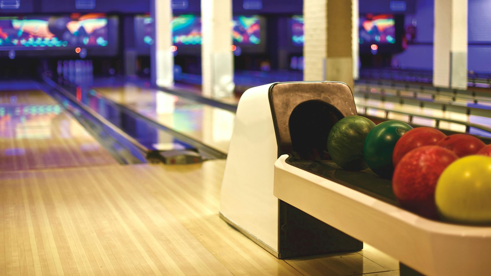 Bowling alley with colorful balls in foreground, lanes extending to background with screens and lighting.