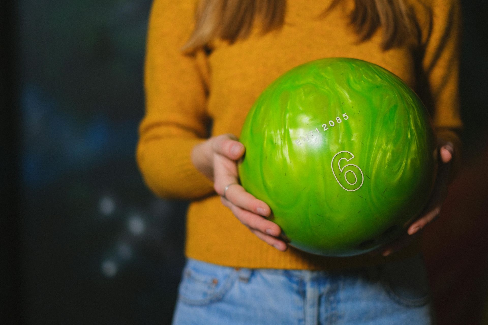 Person holding a green bowling ball with the number 6 on it, against a blurred background.