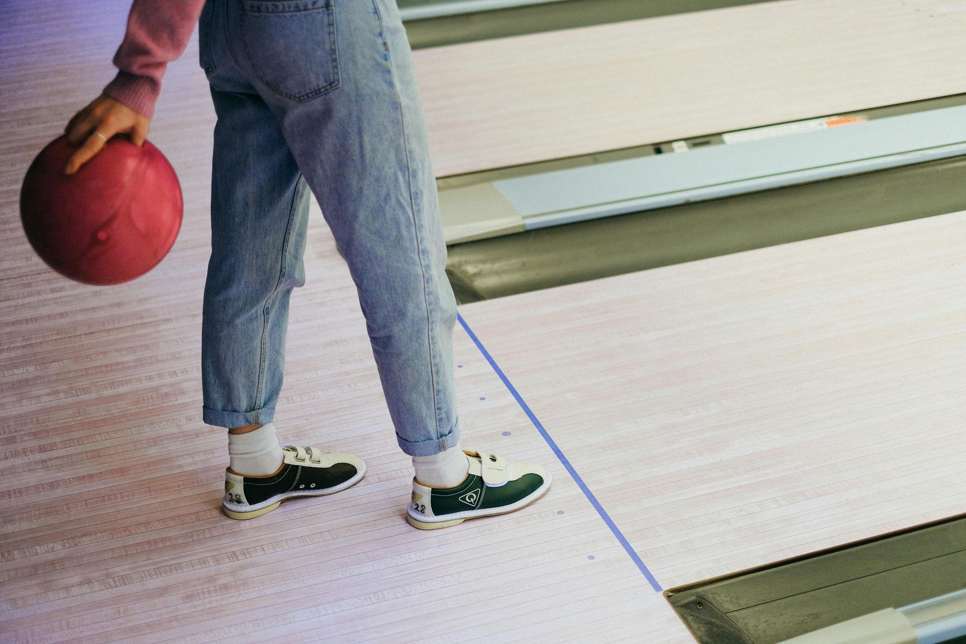 Person in jeans and sneakers holding a red bowling ball, standing on a lane in a bowling alley.
