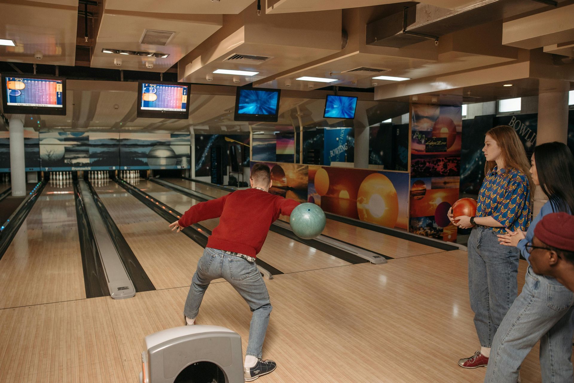 Man bowling, throwing a ball. Three people watch at the lane. Bowling alley interior.