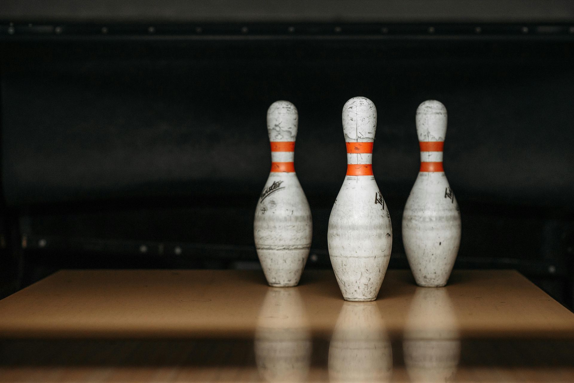 Three bowling pins standing on a wooden lane, in front of a dark backdrop.